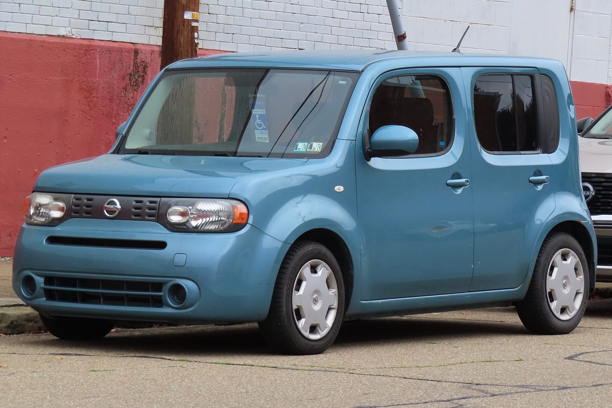 A light blue 2009 Nissan Cube parked on a cracked street in front of a brick wall painted red at the bottom