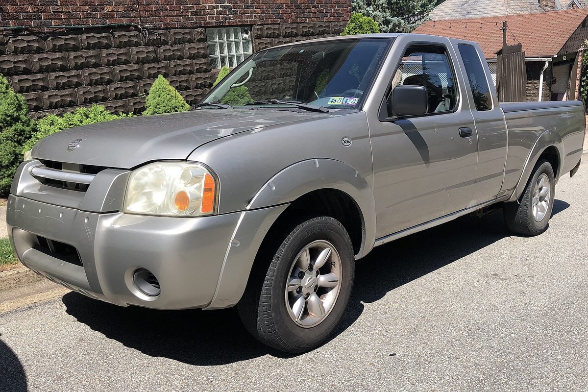 A silver 2004 Nissan Frontier XE King Cab pickup parked at the curb on a residential street, in front of a brick house