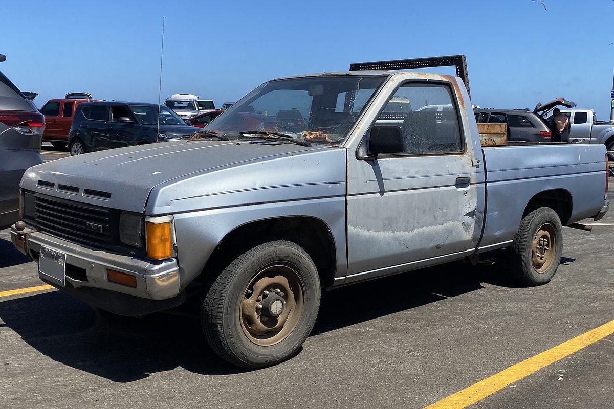 A faded light-blue 1987 Nissan D21 short-bed pickup with surface rust on the hood and roof and a dented door, parked in a sunny lot