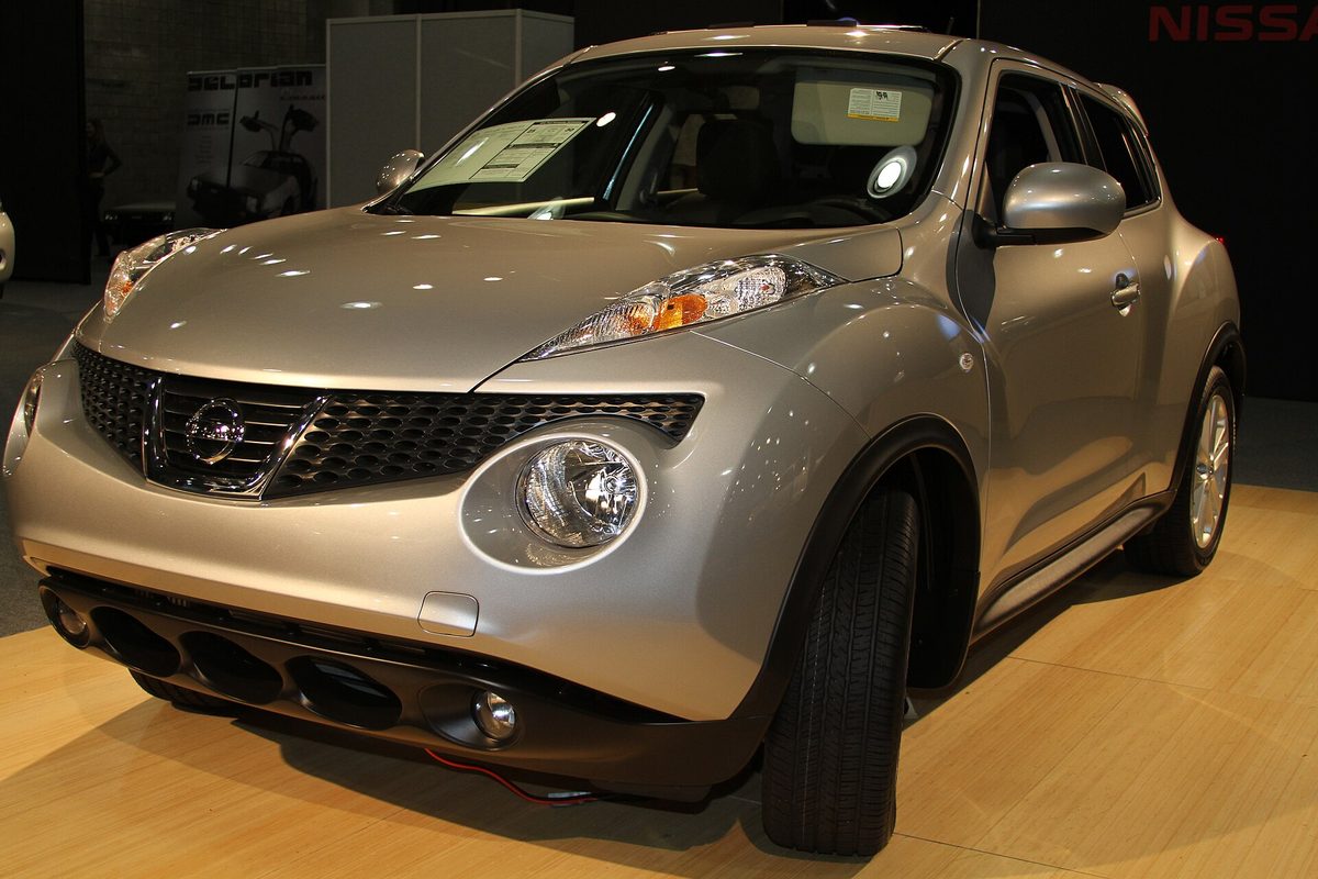 A silver first-generation Nissan Juke on a wooden auto show floor, with a paper sticker inside the windshield