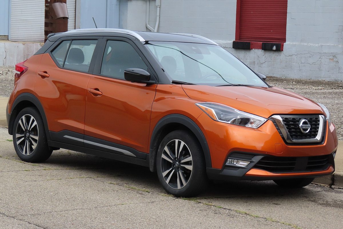An orange 2020 Nissan Kicks SR with a black roof, parked on a cracked concrete lot in front of a gray building