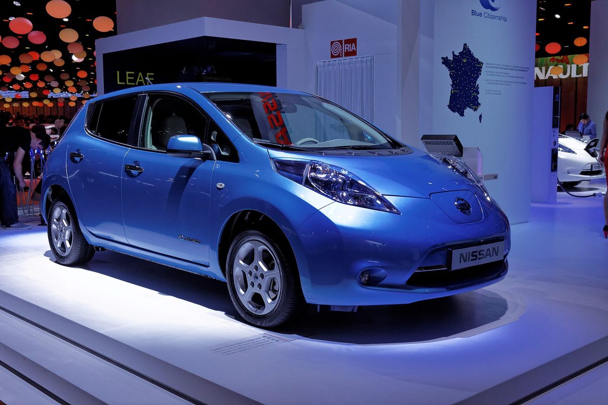 A bright blue first-generation Nissan Leaf hatchback on a lit display stand at the 2012 Paris Motor Show