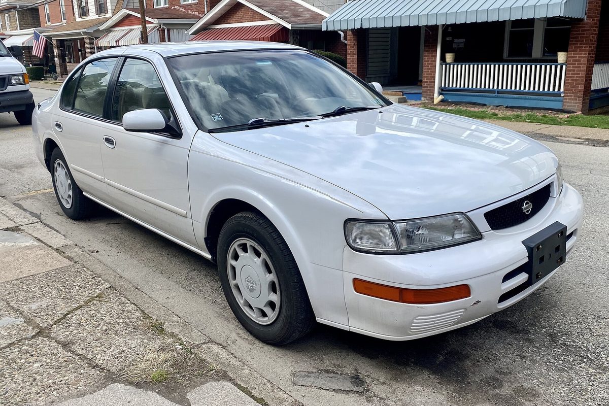 A white 1996 Nissan Maxima GXE sedan parked at the curb on a street of brick row houses with front porches