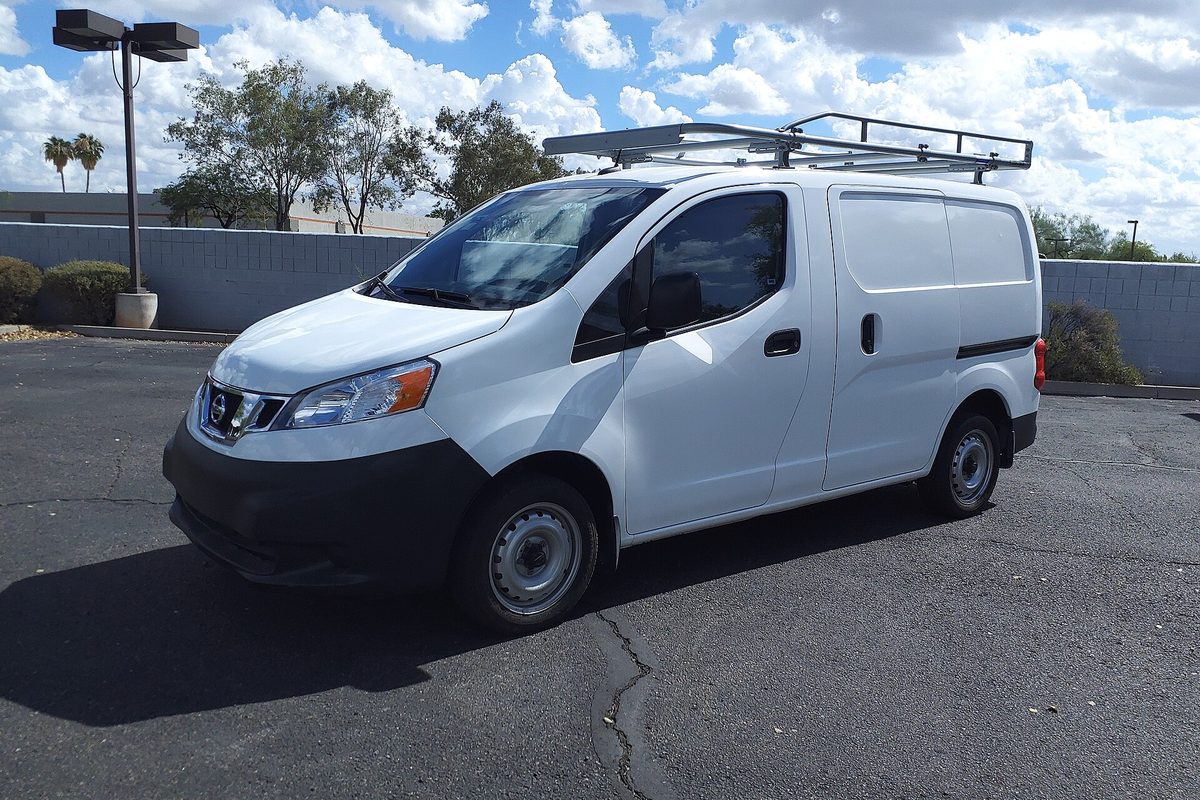 A white 2021 Nissan NV200 S cargo van with a roof ladder rack, parked on an empty lot with palm trees behind