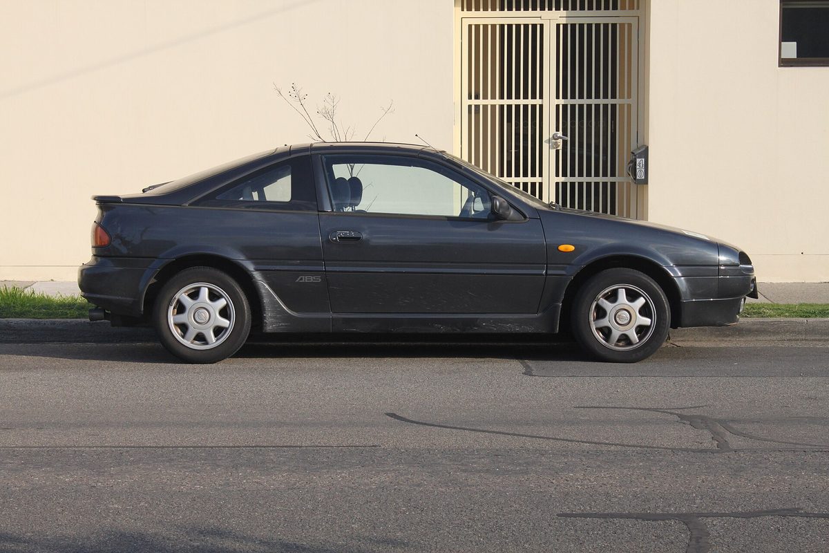 A dark grey 1994 Nissan NX coupe, the same B13 body sold in the US as the NX1600 and NX2000, parked at a curb by a cream wall