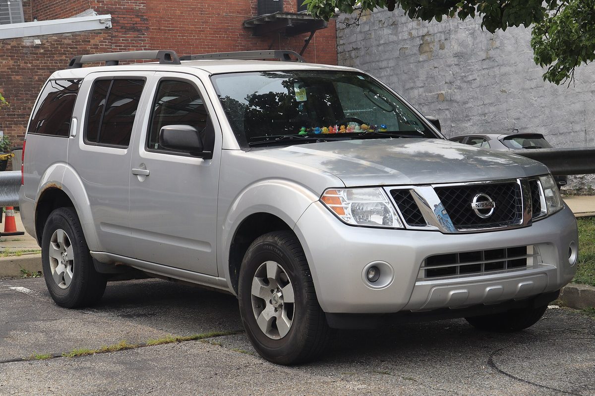 A silver 2011 Nissan Pathfinder S with roof rails, parked on a cracked lot beside a brick building