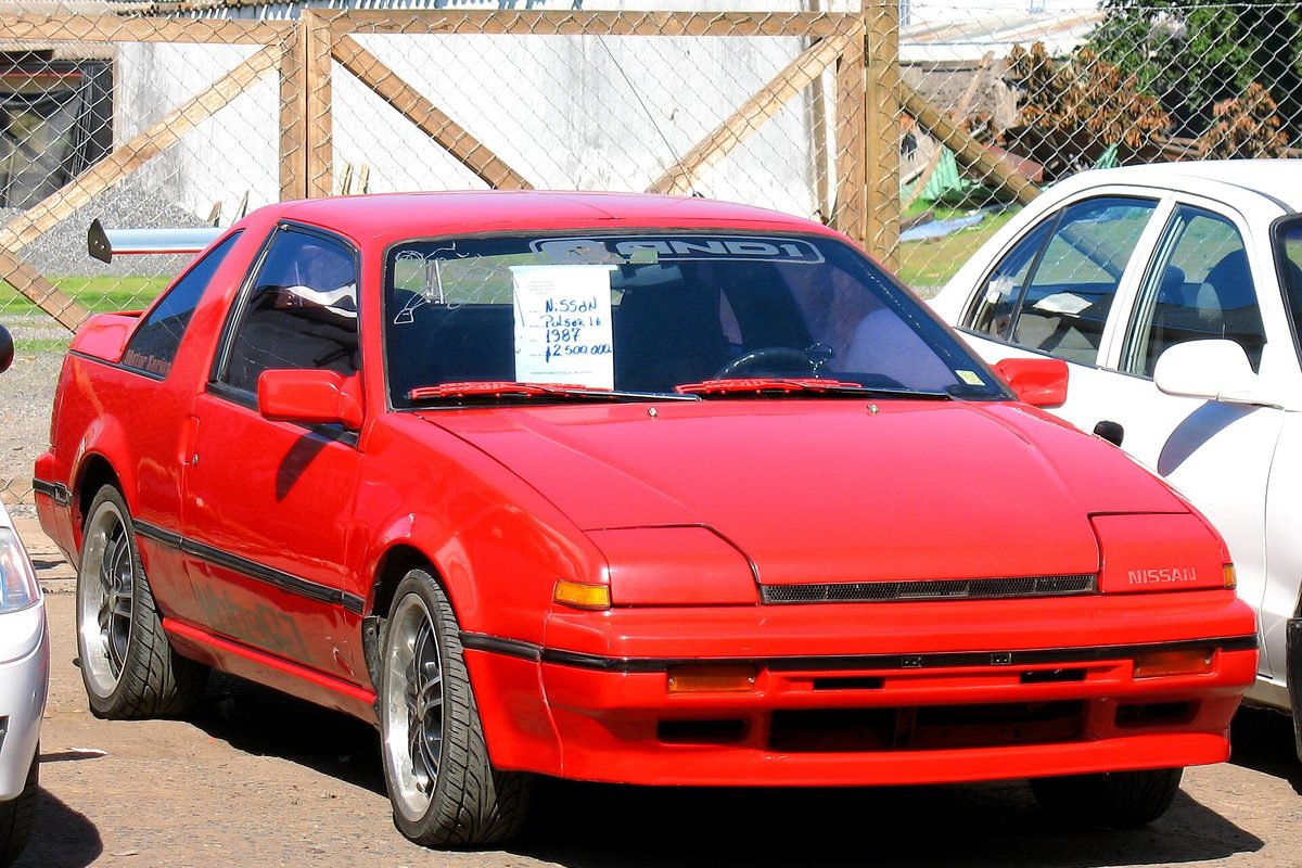 A red 1987 Nissan Pulsar NX coupe with a handwritten sign behind the windshield, parked by a wooden fence and chain-link gate