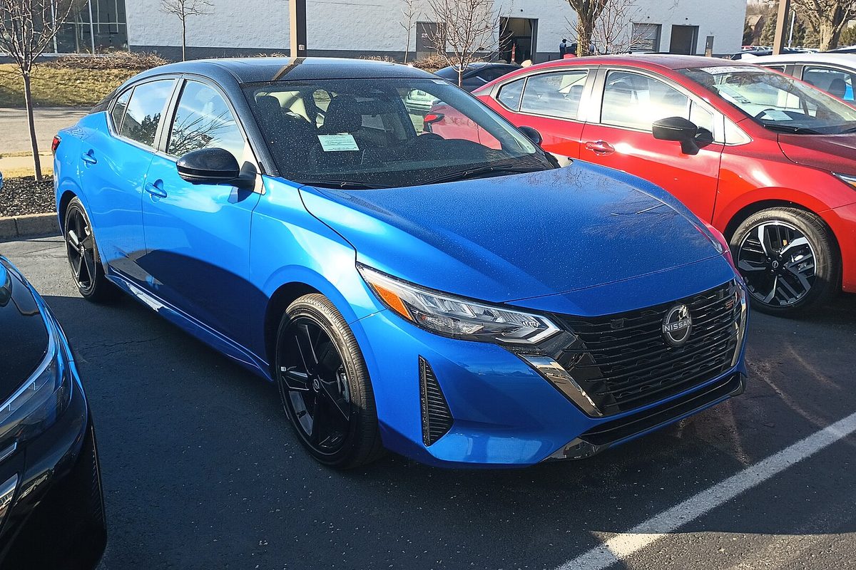 A bright blue 2024 Nissan Sentra SR with black wheels on a dealer lot, a red sedan parked beside it