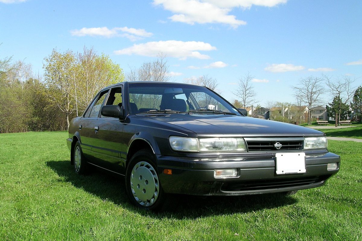 A dark gray 1991 Nissan Stanza sedan parked on a green lawn under a blue sky with a few clouds