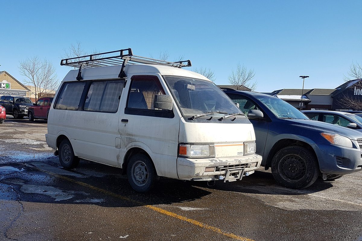 A white Japanese-market Nissan Vanette C22, the van sold in the US as the Nissan Van, with a roof rack on a wet parking lot