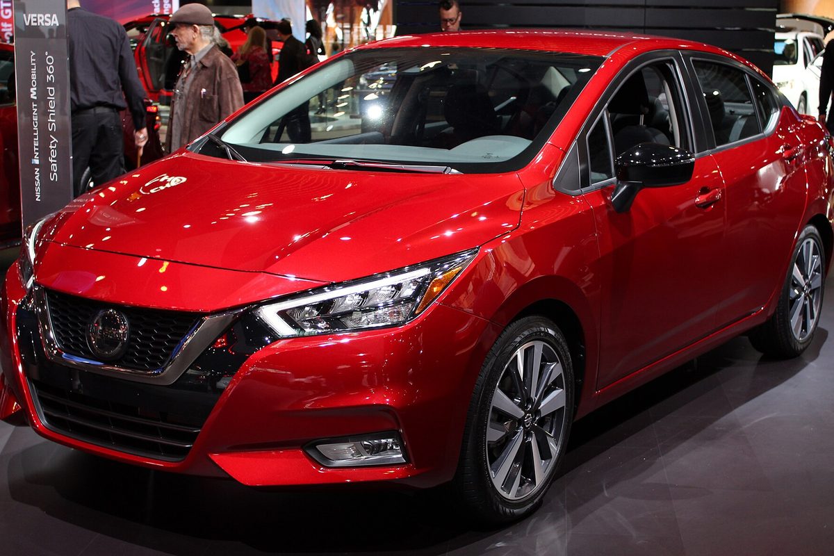 A red 2020 Nissan Versa SR sedan on an auto show floor, with visitors walking behind it
