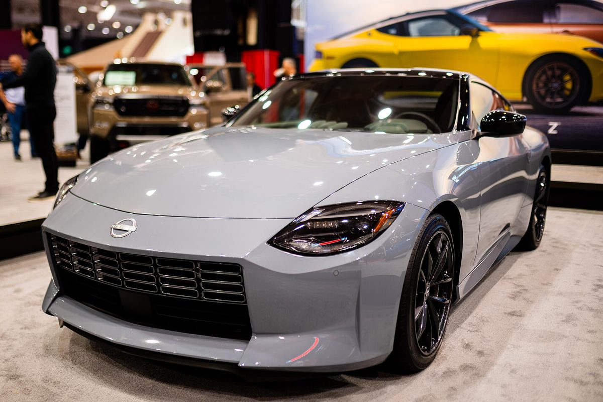 A gray seventh-generation Nissan Z coupe on an auto show stand, with a yellow Z pictured on the display wall behind it