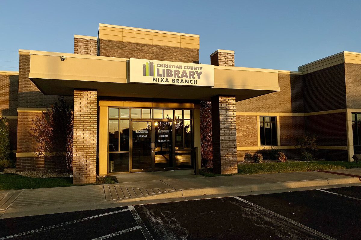 The Christian County Library Nixa Branch, a brick building with a covered glass entrance, lit by low evening sun above its parking lot