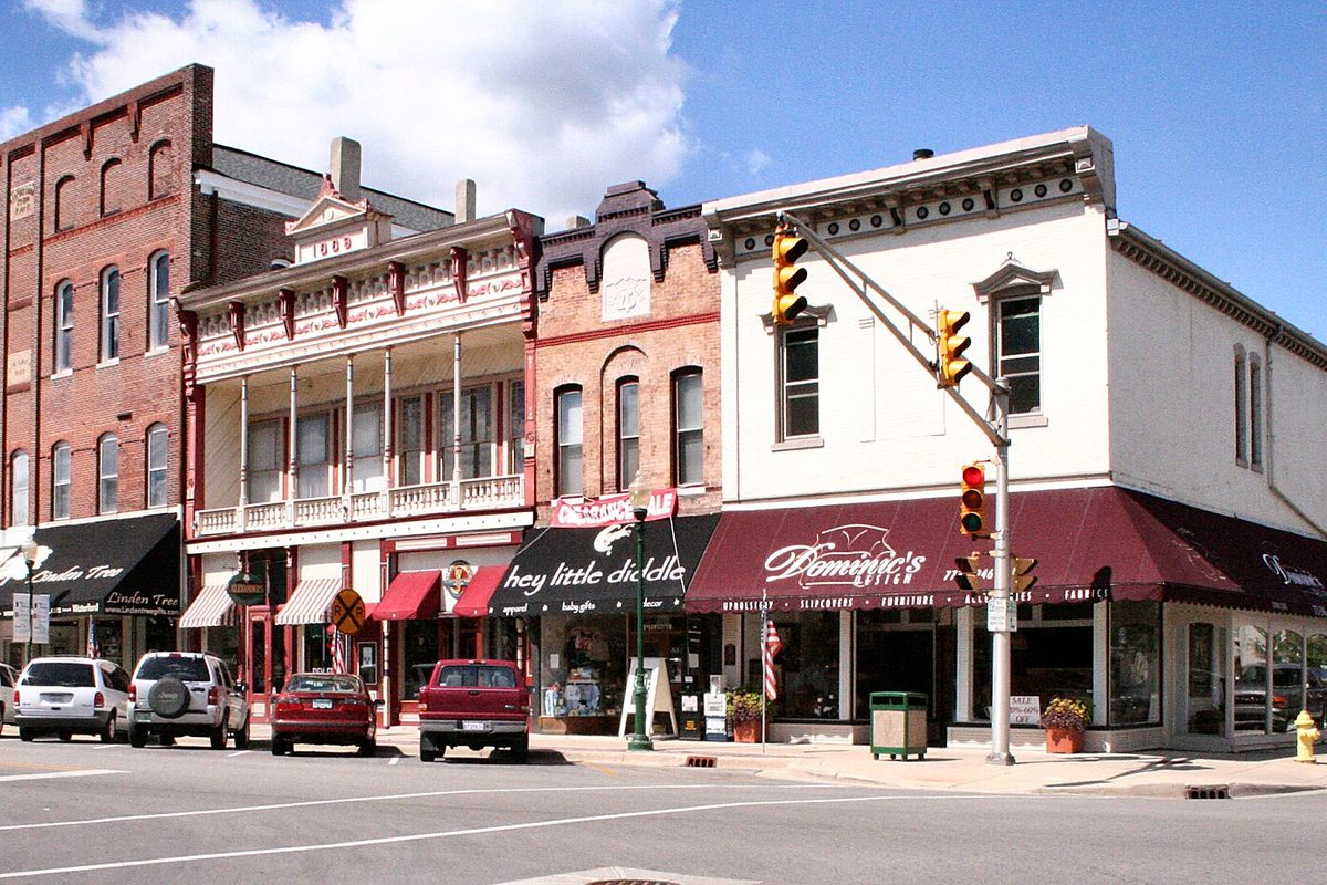 A downtown Noblesville corner of brick and painted storefronts with awnings, traffic lights and cars parked at the curb under a blue sky
