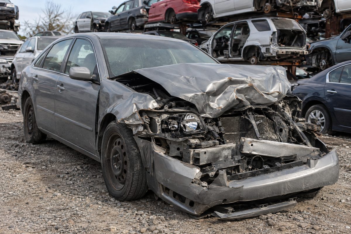 A gray sedan with its front end crushed and the engine bay exposed, sitting in a salvage yard with wrecked cars stacked behind