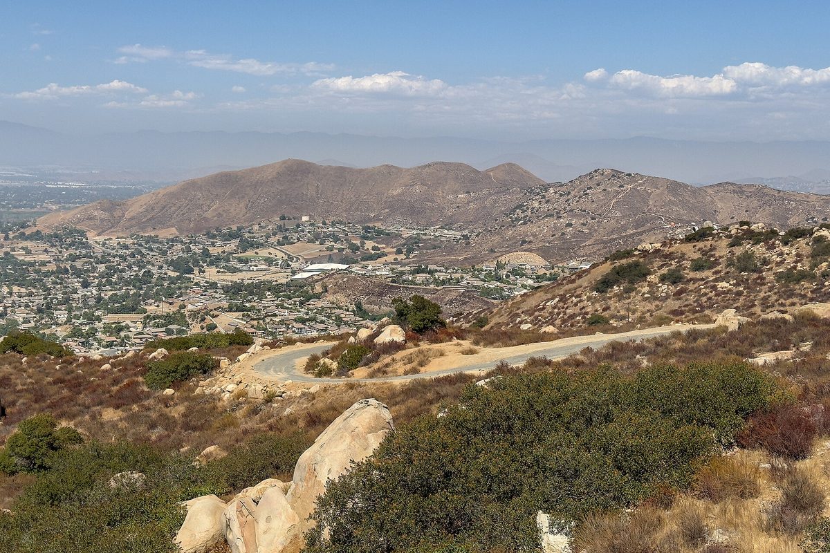 View from the Norco Hills over granite boulders, sage scrub and a dirt trail toward houses and brown hills under a hazy sky