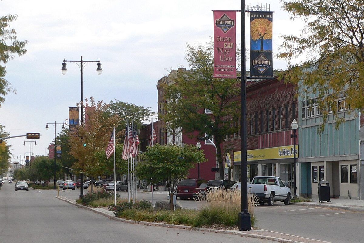 Norfolk Avenue in downtown Norfolk, with brick storefronts, a row of American flags in a planted median, tall banner poles and a white pickup parked at the curb