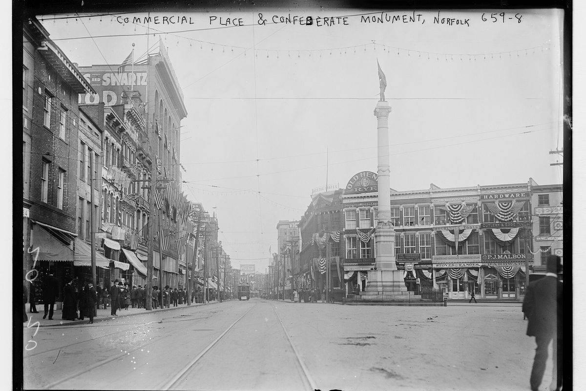 Early 1900s black-and-white photo of Commercial Place in Norfolk: a tall monument column in the square, storefronts hung with flag bunting, and streetcar tracks in the road