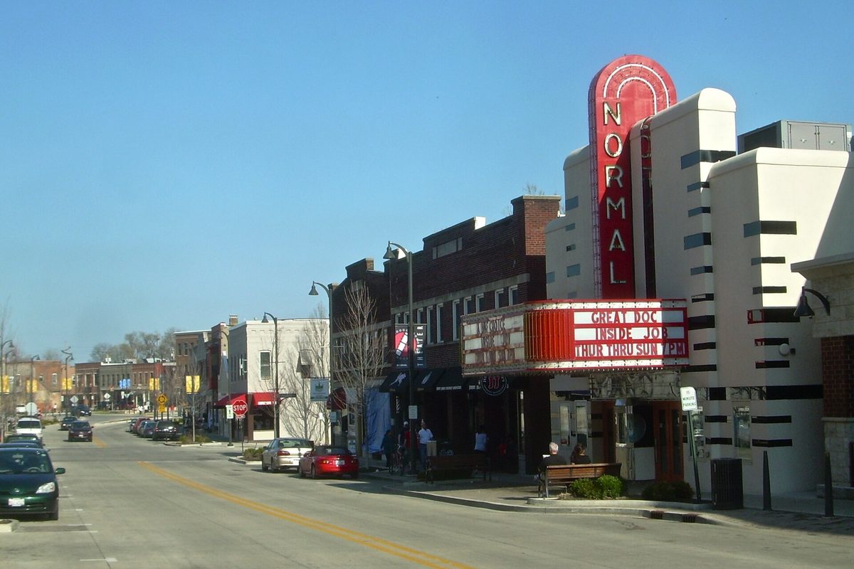 North Street in Normal looking east, with the red vertical sign and marquee of the Normal Theater beside brick storefronts and parked cars