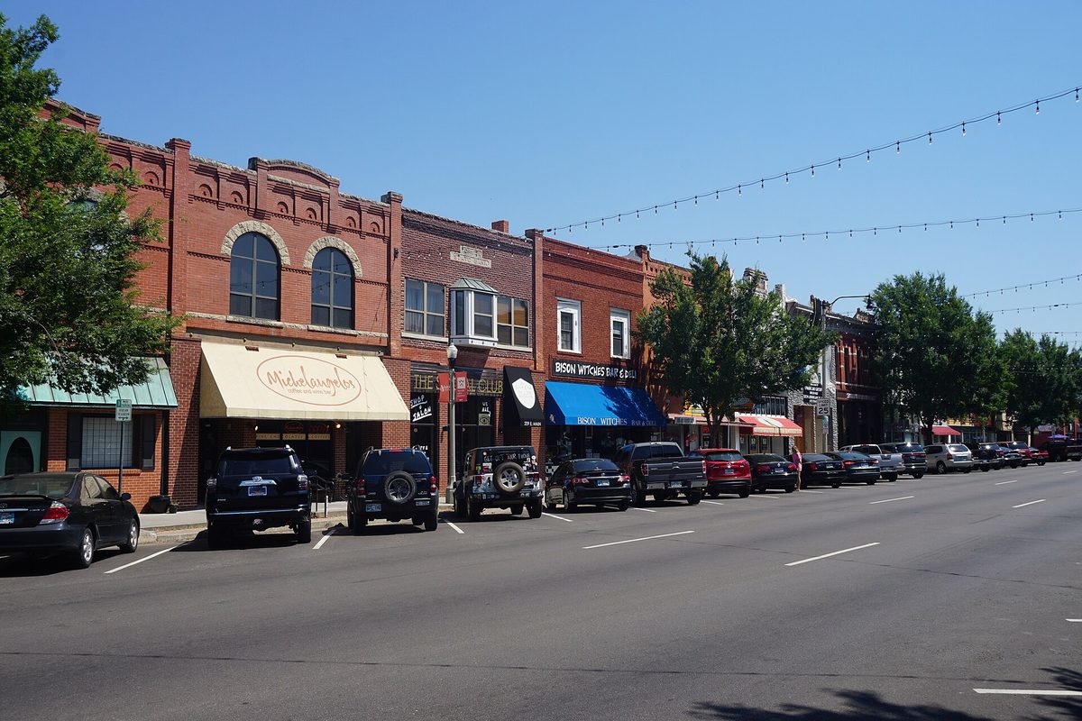 A row of brick storefronts with awnings on East Main Street in Norman, cars parked at the curb under strings of lights and a clear blue sky