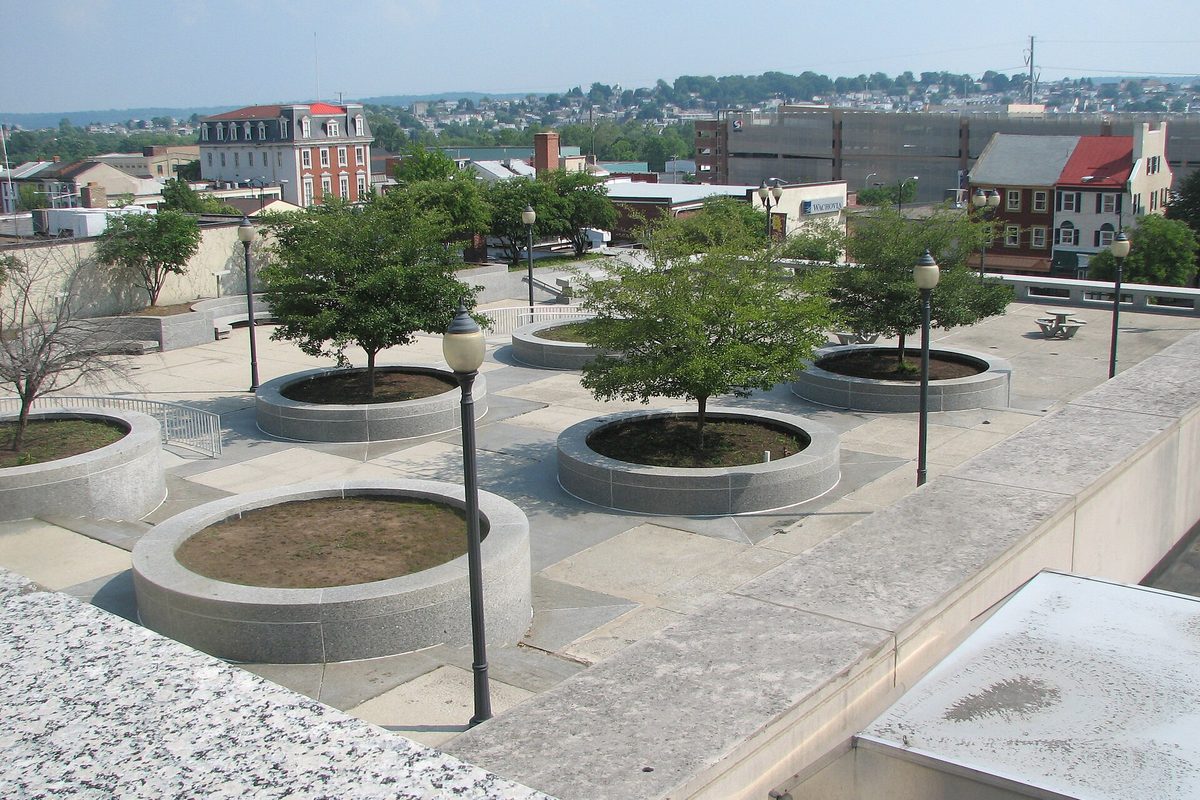 A paved rooftop plaza with round stone tree planters and black lampposts, looking out over brick buildings in central Norristown toward wooded hills