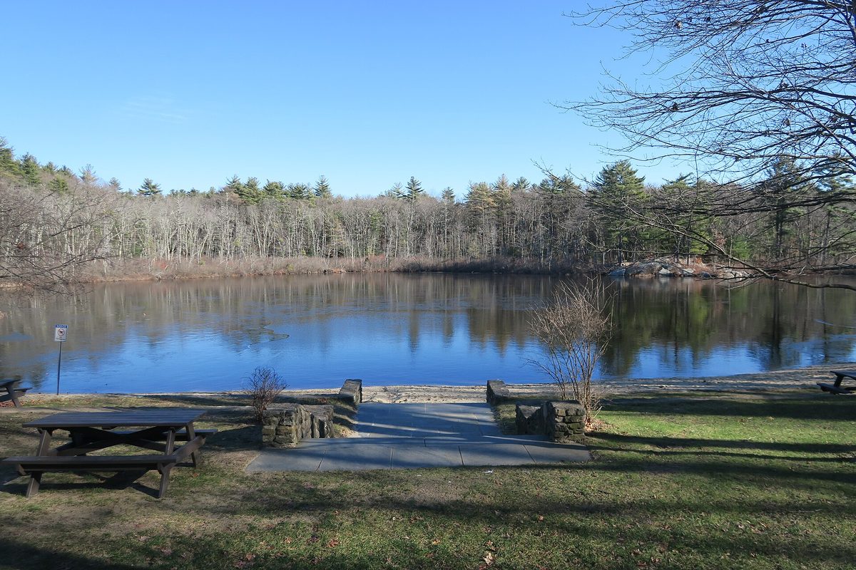 Berry Pond in Harold Parker State Forest, North Andover, with bare trees and pines reflected in still blue water and a picnic table on the grass in front
