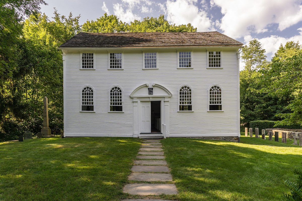 The white clapboard Old Narragansett Church in Wickford, with arched windows and a stone path leading to its open door, old gravestones at its side