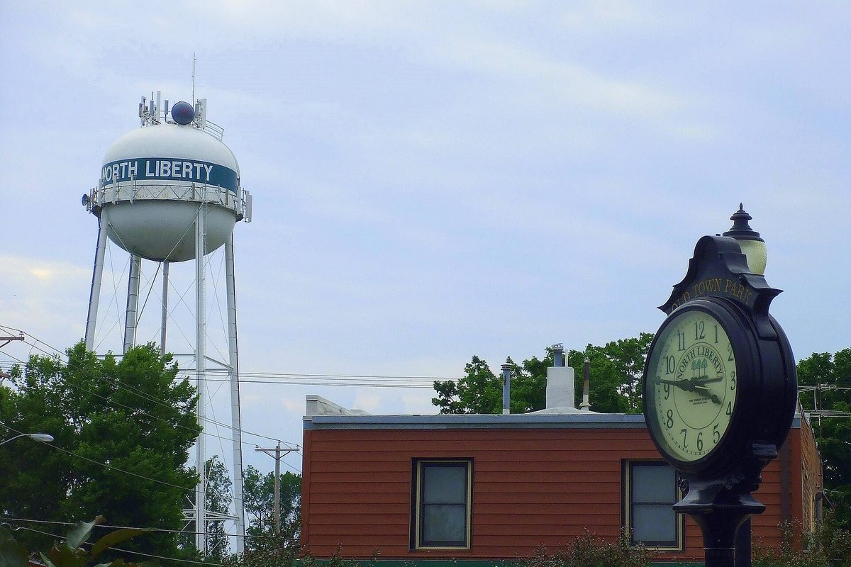 The white North Liberty water tower behind a black street clock and a low red building under a gray sky