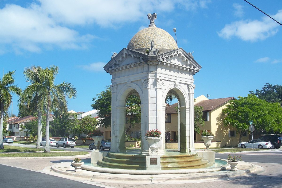 The Fulford by the Sea entrance monument in North Miami Beach, a white stone pavilion with arches and a dome over a fountain in a traffic circle, with palms and homes behind