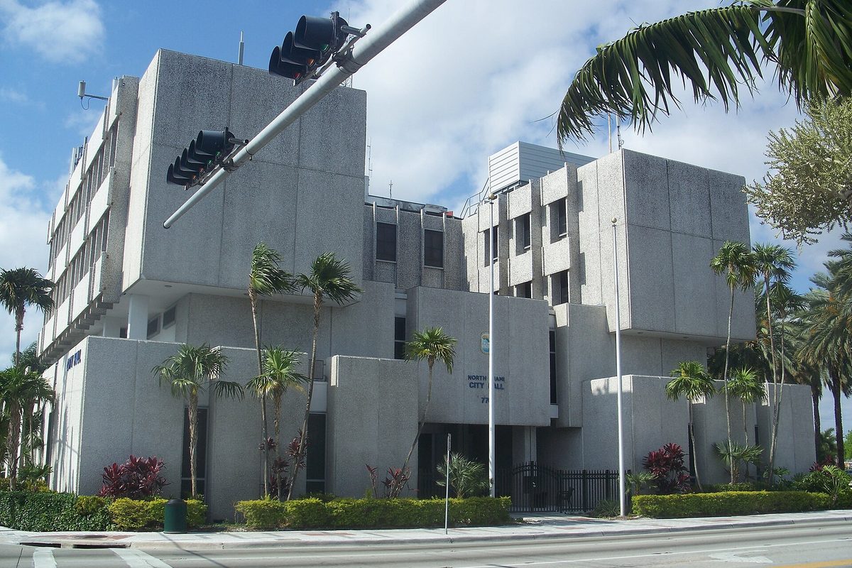 North Miami City Hall, a stacked concrete building with deep-set windows, palms and red-leaf shrubs out front and a traffic signal overhead