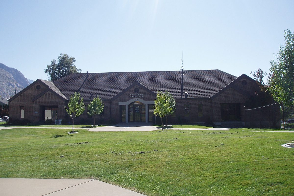 North Ogden's low brick city hall with a dark shingled roof behind a wide green lawn, rocky mountain slopes rising at the left