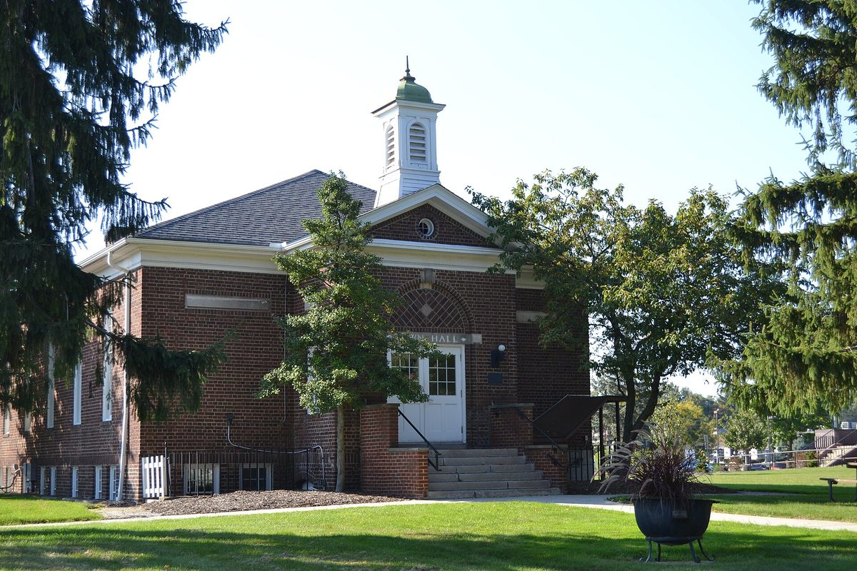 North Olmsted's old town hall, a one-story red-brick building with a white cupola and a green roof cap, behind a lawn and tall evergreens