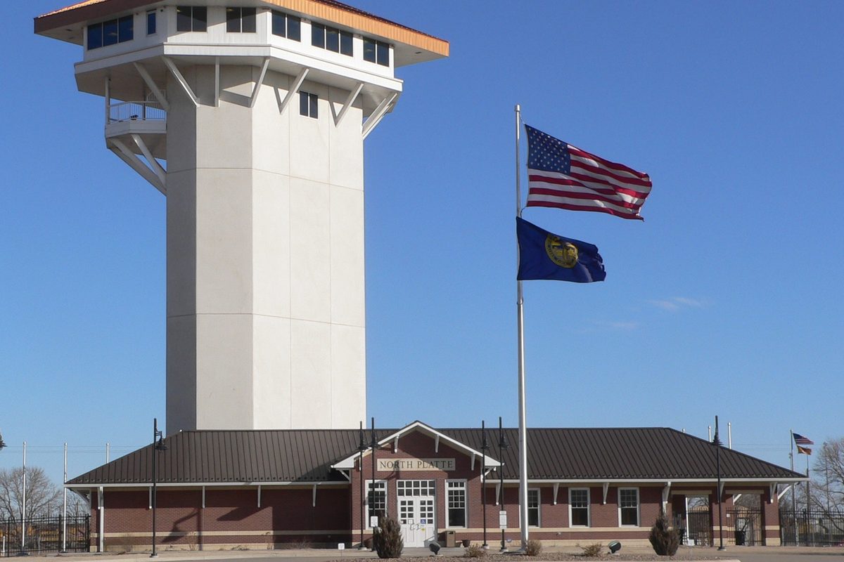 The white Golden Spike Tower rising behind a red-brick visitor center with North Platte over the door, with the US and Nebraska flags flying out front under a blue sky