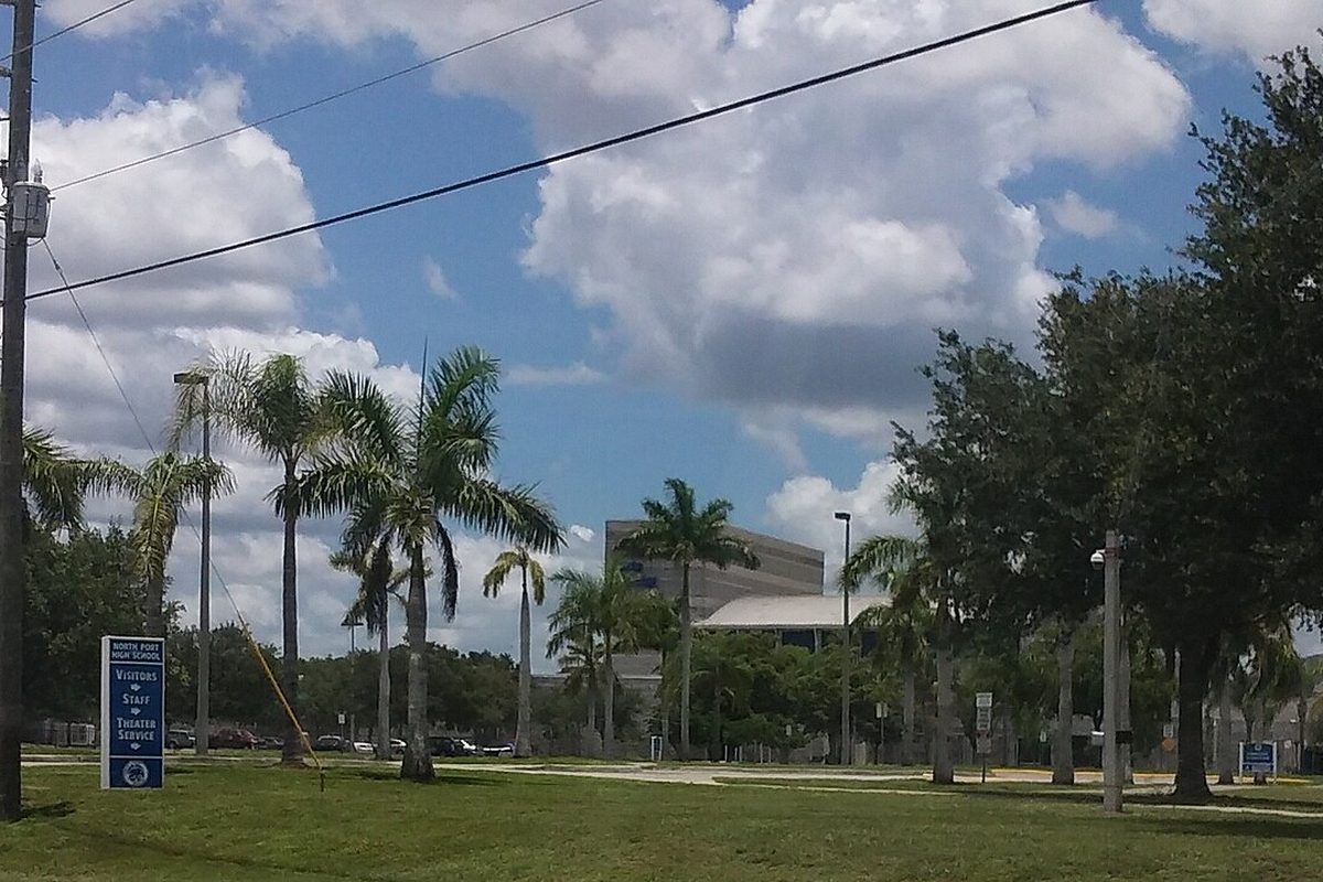 The entrance to North Port High School: a blue visitor sign, a wide lawn, tall palm trees and a school building under a cloudy sky