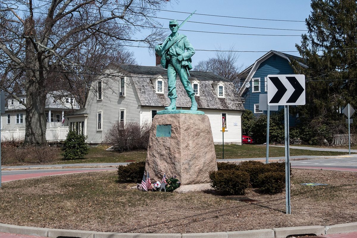 The green bronze Soldiers and Sailors Monument, a Civil War soldier holding a rifle, on a granite base in the traffic circle at Fruit Hill and Olney Avenues, with a white house behind it