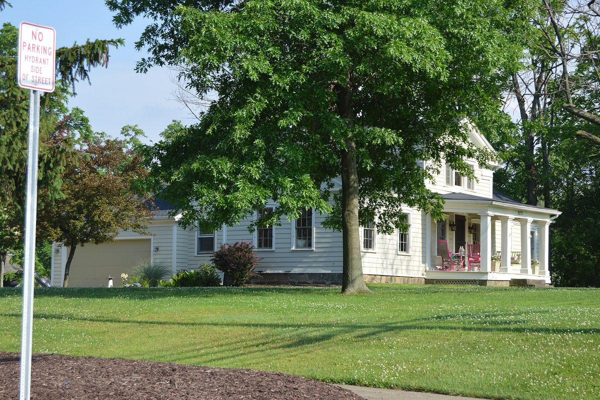 A white two-story farmhouse with a columned front porch behind a wide lawn and a large shade tree, with a No Parking Hydrant Side of Street sign in the foreground