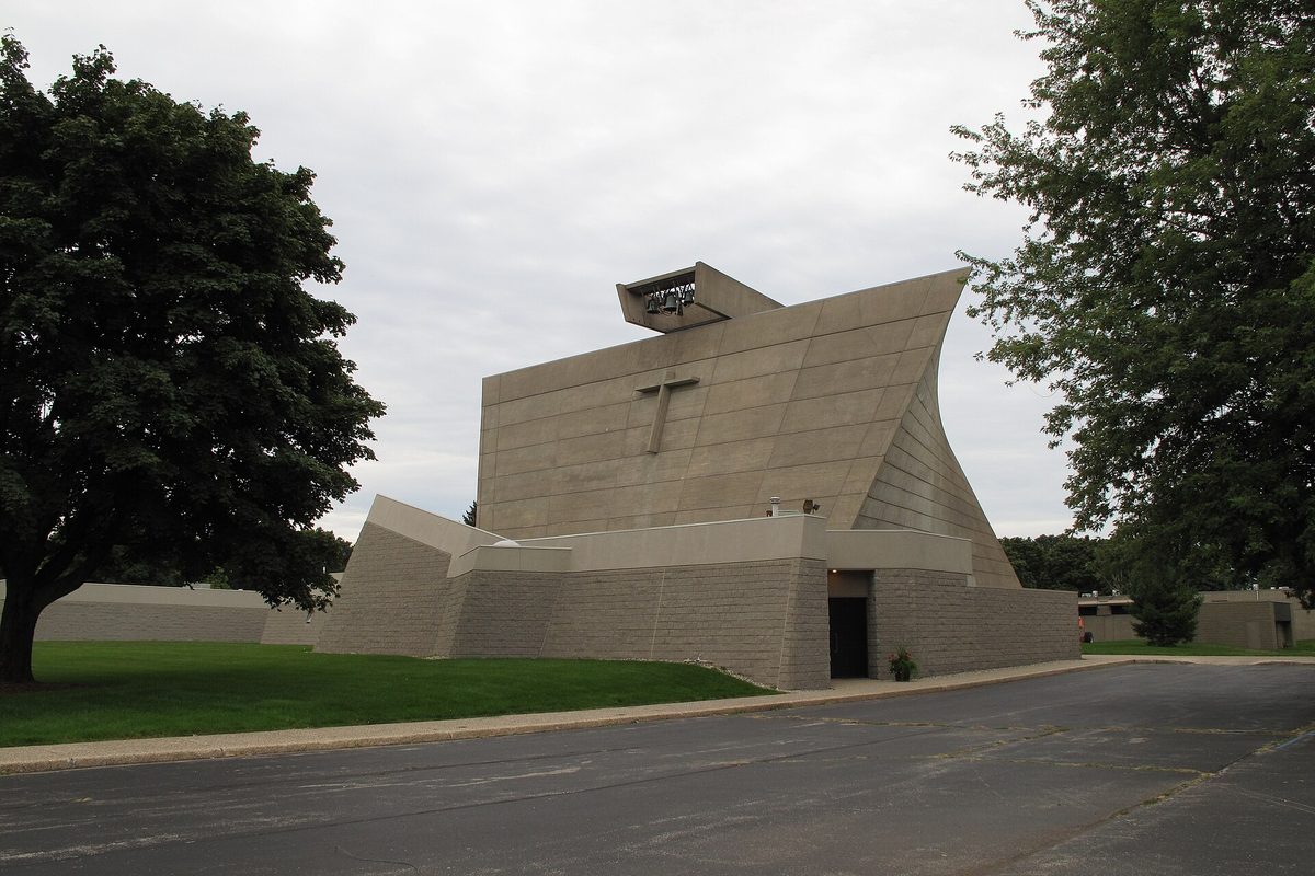 St. Francis de Sales Church in Norton Shores, a tall slanted concrete building with a cross on its face, beside a large tree and an empty parking lot