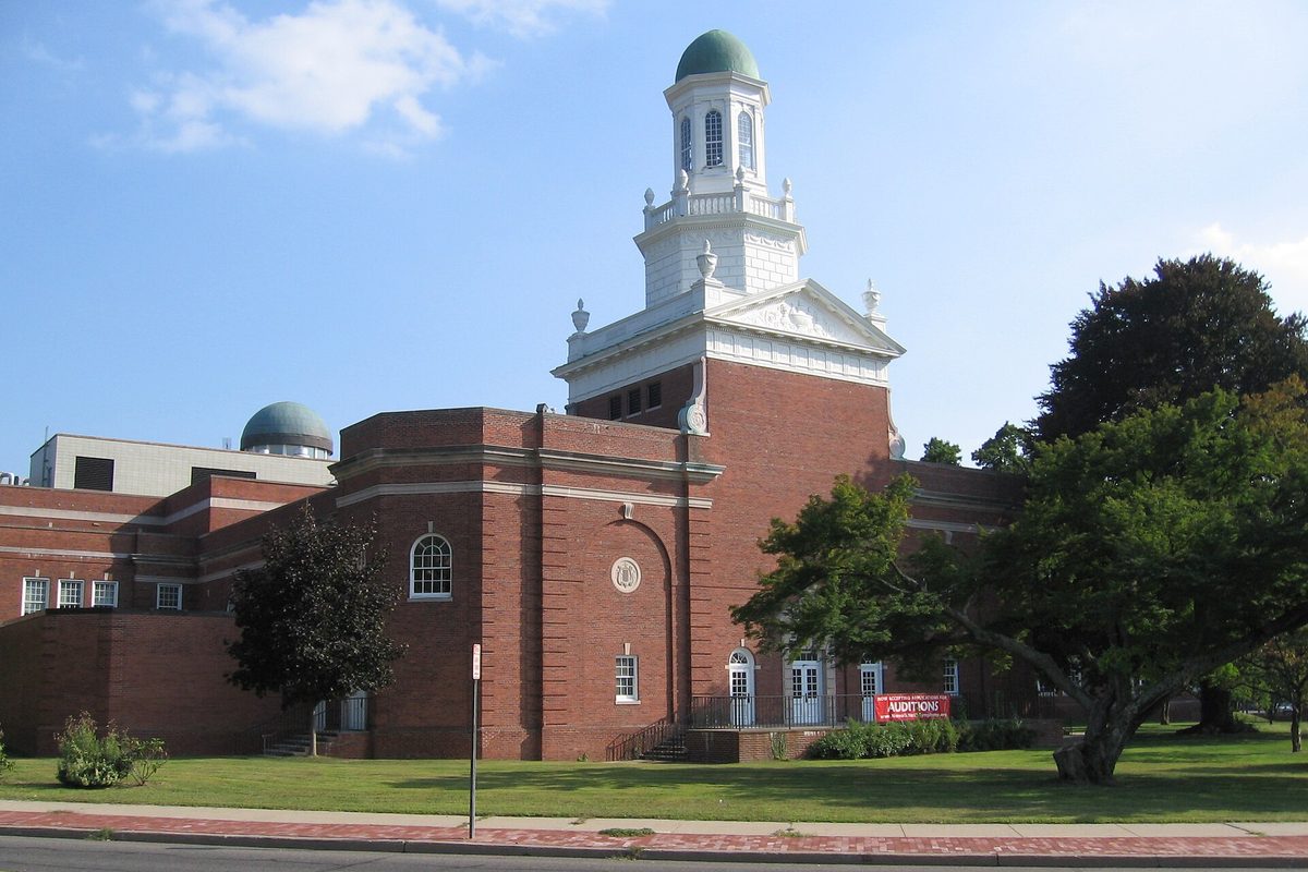 Norwalk City Hall, a red brick building with a white tower and green dome, behind a green lawn and brick sidewalk on a sunny day