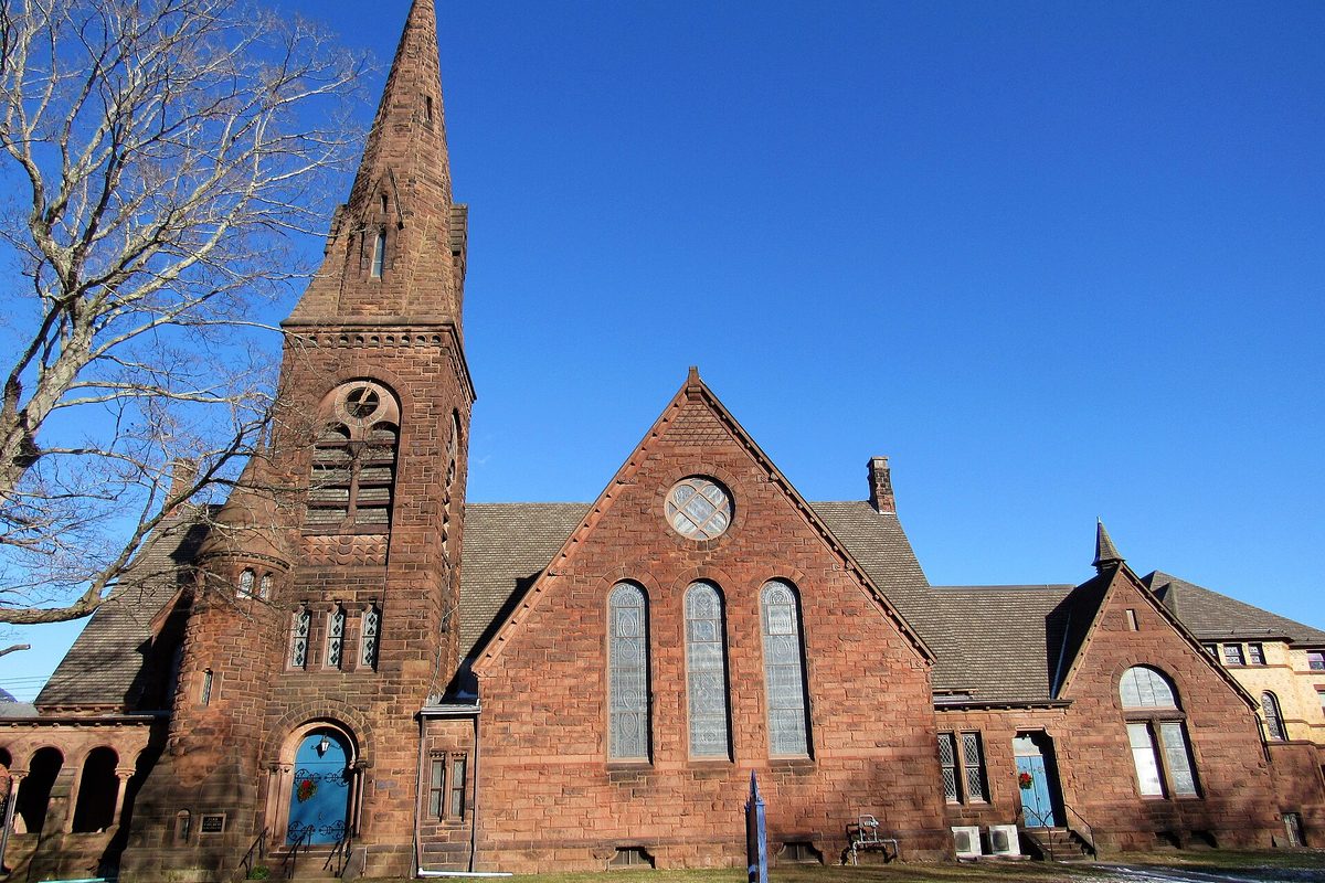 Park Congregational Church in Norwich, a reddish-brown stone church with a tall spire, a round window and arched blue doors, under a clear blue sky