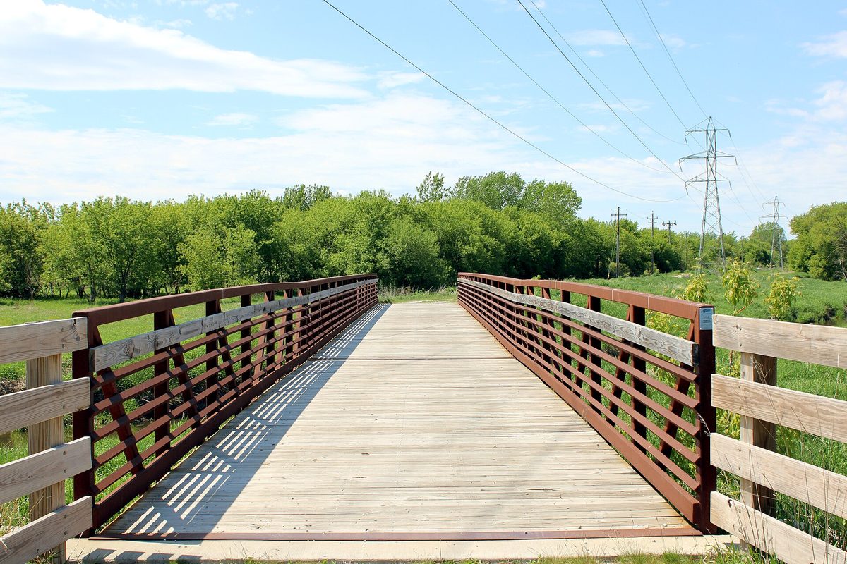 A wooden pedestrian bridge with rust-red steel railings on the Oak Leaf Trail in Oak Creek, with green trees and power lines beyond