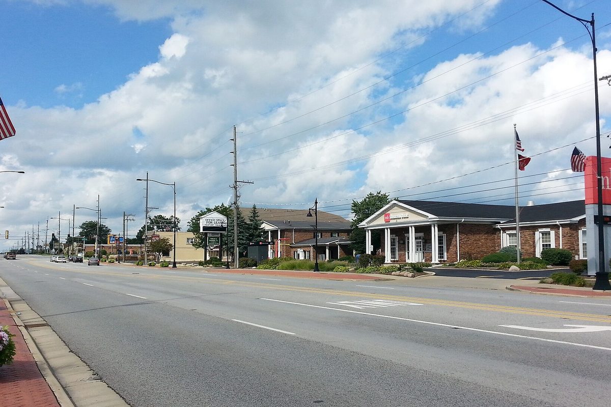 Cicero Avenue in Oak Forest, a wide road lined with low brick offices, flagpoles and streetlights under a cloudy blue sky