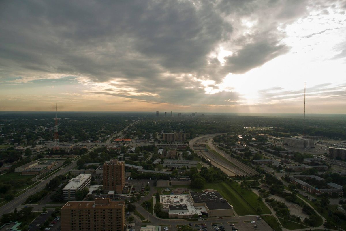 Drone view over Oak Park at sunset, with Interstate 696 curving through, apartment towers in the foreground and low buildings stretching to the horizon under heavy clouds