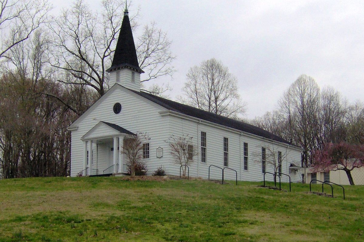 The white wooden Chapel-on-the-Hill in Oak Ridge, with a black steeple and columned porch, on a grassy rise among bare trees
