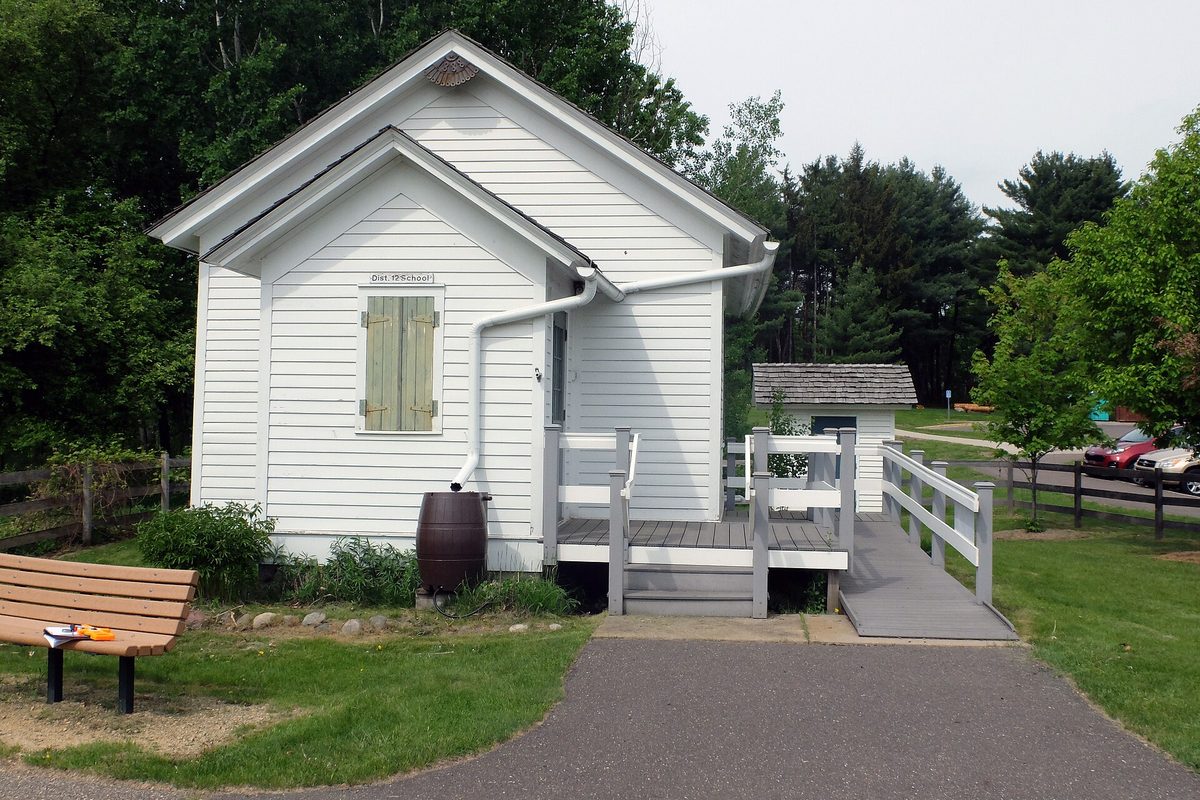 The white clapboard Eder Schoolhouse, built in 1888, with green shutters, a rain barrel and a wooden ramp at the Oakdale Nature Center