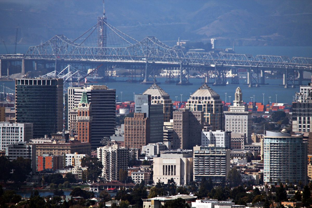 Downtown Oakland towers seen from the hills, with the Bay Bridge and the port's container cranes across the water behind them