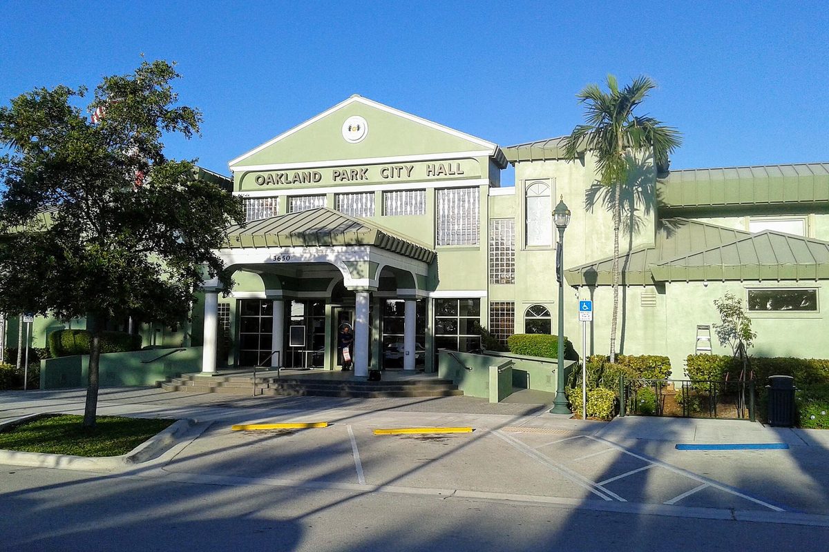 The pale green Oakland Park City Hall with its columned entrance, palm trees and an empty parking area in late-day sun