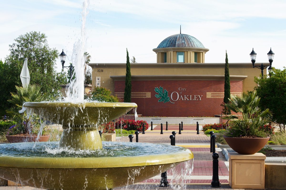 A tiered fountain in front of Oakley City Hall, a tan building with a teal dome and a red wall bearing the City of Oakley sign