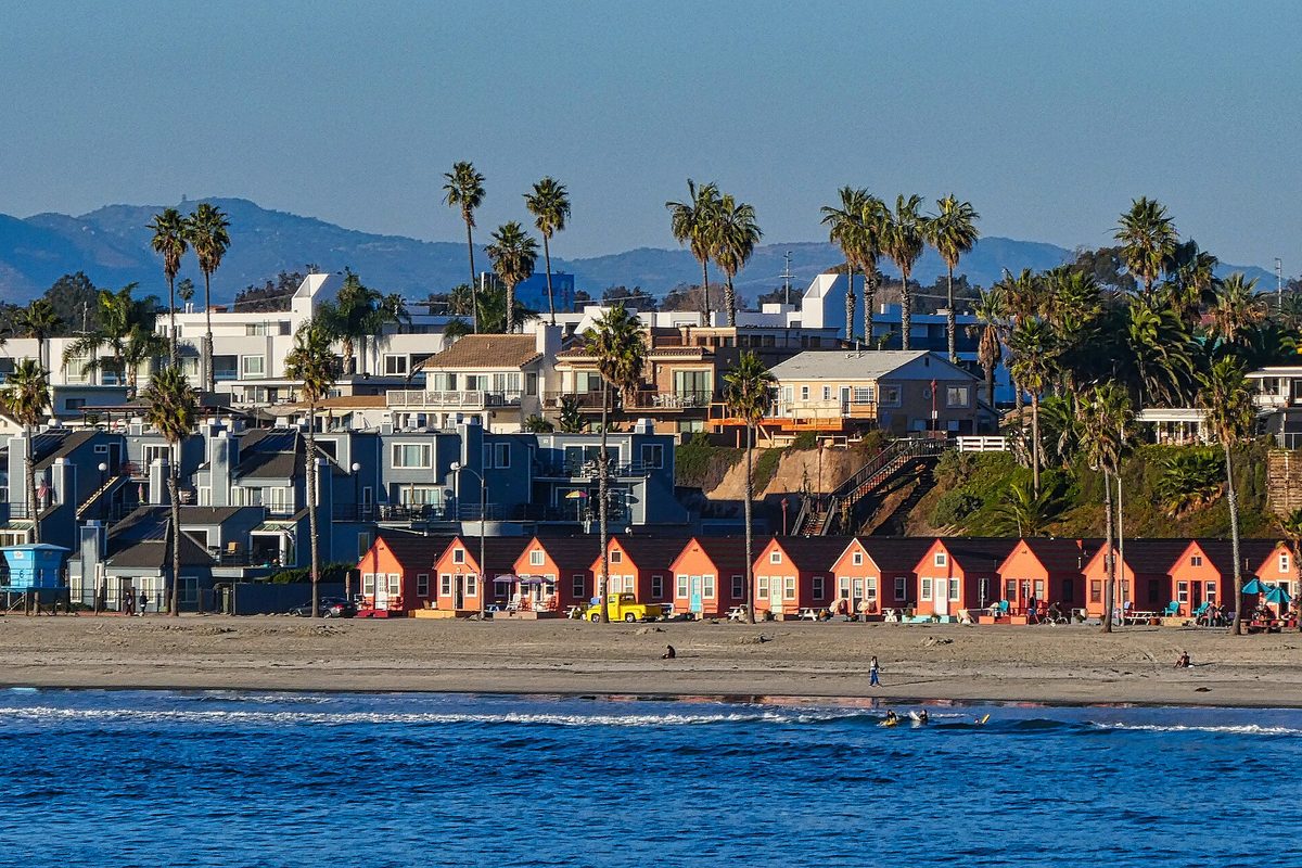 Oceanside's beach seen from the pier, with a row of orange-red beach cottages, palm trees and hillside homes below distant mountains