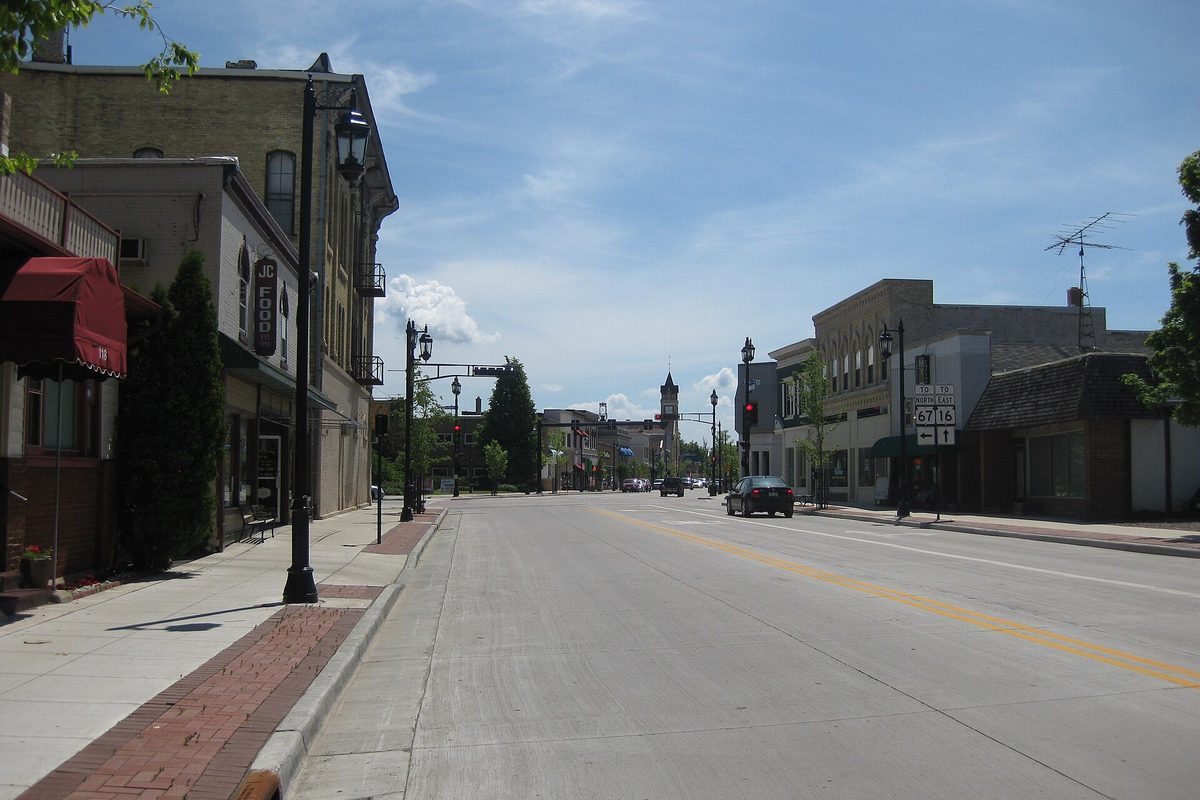 A wide, nearly empty main street in downtown Oconomowoc on a sunny day, lined with two-story storefronts, black lamp posts and a clock tower in the distance