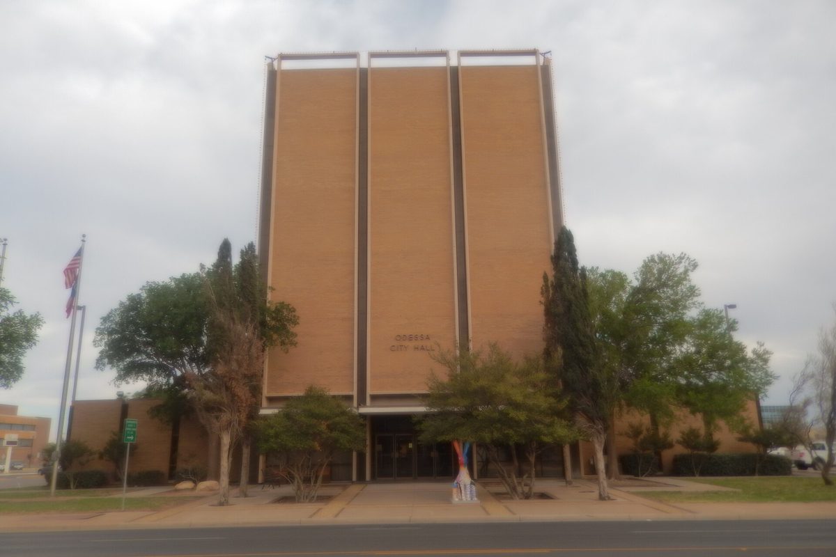 Odessa City Hall, a tall tan tower with narrow vertical panels, behind trees and an American flag under a gray sky